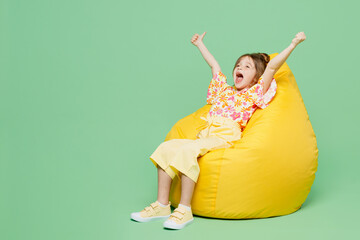 Wall Mural - Full body excited winner little child kid girl 6-7 years old wears casual clothes sit in bag chair show thumb up isolated on plain pastel green background. Mother's Day love family lifestyle concept.