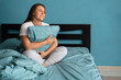 © Andrii Lysenko - Young woman hugging blue pillow sitting on comfortable bed in the morning