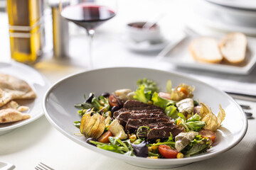Wall Mural - Beef steak sliced on top of mixed salad with greens, physalis, cherry tomatoes served with wine
