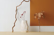 © TrueFrame Collective - Horizontal shot of contemporary still life installation with gypsum vessel, dried flowers, monstera leaf and branch against white and terracotta brown wall background, copy space