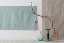 Horizontal Shot Of Minimalistic Still Life Installation With Glassware, Branch And Dried Red Flowers Against White Wall, Gray And Blue Linen Fabric Background, Copy Space