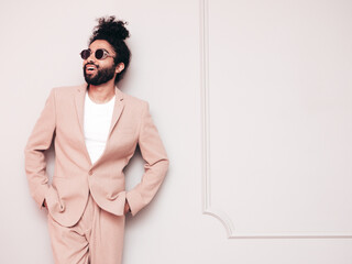 Handsome smiling hipster  model. Unshaven Arabian man dressed in elegant beige suit . Fashion male with long curly hairstyle posing near grey wall in studio. Isolated. In sunglasses