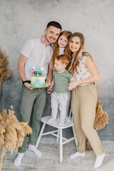 Happy family holds cake and having fun celebrating a birthday party 2 years. Mom, dad, daughter congratulate son on birthday near grey wall in studio. Mother, father hugging kids. Birthday reception.