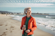 Smiling Mature Woman In Green Jacket And Red Jacket On The Beach With Her Mobile Phone And Headphones Listening To Music Image Ai Generate