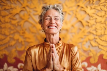 Wall Mural - Close-up portrait photography of a happy mature woman joining palms in a gesture of gratitude against a gold background. With generative AI technology
