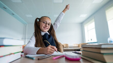 Caucasian Girl With Two Ponytails Sits At Her Desk And Raises Her Hand Up To Answer The Teacher's Question.