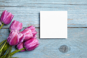 Card mockup, white blank wedding invitation with floral decor on bue wooden background. Square greeting card mockup with purple flowers on table