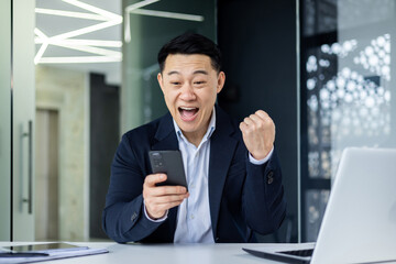 Happy young Asian man working in the office, sitting at the table and using the phone, looking at the screen, showing a victory gesture with his hand, happy, celebrating.