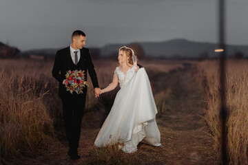 Wall Mural - Beautiful bride and groom in evening lights, walking and hugging holding a bouquet of flowers