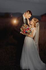 Wall Mural - Beautiful bride and groom in evening lights, kissing and hugging holding a bouquet of flowers