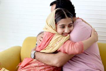 Wall Mural - Indian Muslim daughter girl with traditional hijab scarf sitting with her father in living room, child hugging her dad, smiling cute kid spending time together with daddy at home, happy lovely family.