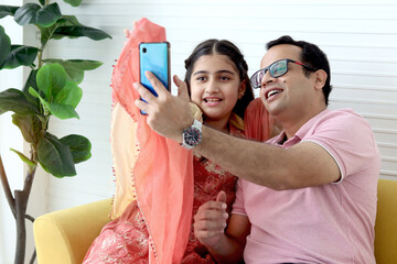 Wall Mural - Indian happy family spending time together, cute daughter girl kid in India traditional dress costume and her father taking a selfie by mobile phone while sitting in living room or making video call.