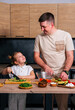 © dvulikaia - Cute little girl and her handsome young dad are cutting vegetables and smiling while cooking in kitchen at home.