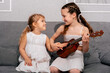 © dvulikaia - Two girls are sitting on the couch with a ukulele. The older sister teaches the younger to play the guitar.