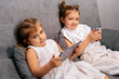 © dvulikaia - Two girls sitting on sofa using modern gadgets to watch videos and play games.