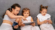 © dvulikaia - Cute little sisters sitting on sofa using modern gadgets to watch videos and play games.