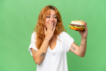 Wall Mural - Young caucasian woman holding a burger isolated on green screen chroma key background with surprise and shocked facial expression