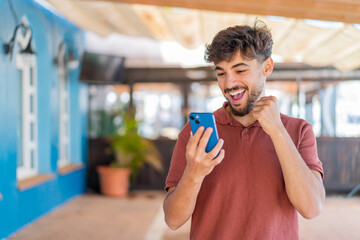 Wall Mural - Young Arabian handsome man at outdoors using mobile phone and doing victory gesture