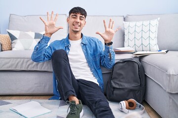 Wall Mural - Young hispanic man sitting on the floor studying for university showing and pointing up with fingers number ten while smiling confident and happy.