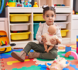 Adorable hispanic girl sitting on floor playing with baby doll at kindergarten