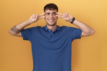 Poster - Young hispanic man standing over yellow background doing peace symbol with fingers over face, smiling cheerful showing victory