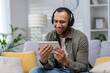 © Tetiana - Young smiling hispanic man sitting on sofa at home wearing headphones and using tablet