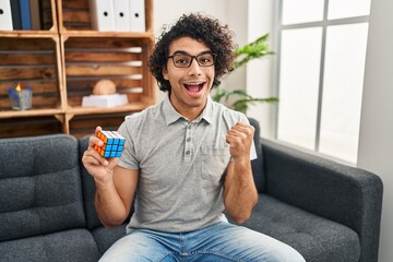 Sticker - Hispanic man with curly hair playing colorful puzzle cube intelligence game screaming proud, celebrating victory and success very excited with raised arms