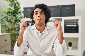 Sticker - Hispanic man with curly hair holding contact lenses making fish face with mouth and squinting eyes, crazy and comical.