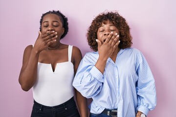 Wall Mural - Two african women standing over pink background bored yawning tired covering mouth with hand. restless and sleepiness.