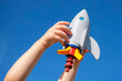 Child arms holding up a plastic toy rocket spaceship against blue sky