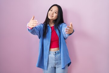 Sticker - Young asian woman standing over pink background looking at the camera smiling with open arms for hug. cheerful expression embracing happiness.