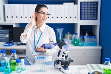 Poster - Young brunette woman working at scientist laboratory smiling with hand over ear listening an hearing to rumor or gossip. deafness concept.