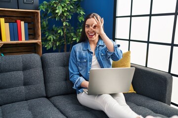 Wall Mural - Hispanic woman using laptop at home smiling happy doing ok sign with hand on eye looking through fingers