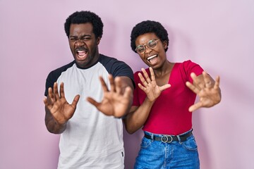 Wall Mural - Young african american couple standing over pink background afraid and terrified with fear expression stop gesture with hands, shouting in shock. panic concept.