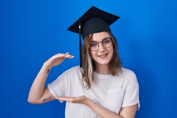 Wall Mural - Blonde caucasian woman wearing graduation cap gesturing with hands showing big and large size sign, measure symbol. smiling looking at the camera. measuring concept.