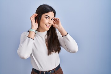 Canvas Print - Young brunette woman standing over blue background trying to hear both hands on ear gesture, curious for gossip. hearing problem, deaf