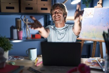 Sticker - Middle age man sitting at art studio with laptop at night looking at the camera smiling with open arms for hug. cheerful expression embracing happiness.