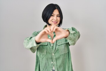 Wall Mural - Young asian woman standing over white background smiling in love doing heart symbol shape with hands. romantic concept.