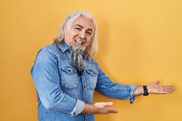 Wall Mural - Middle age man with grey hair standing over yellow background inviting to enter smiling natural with open hand