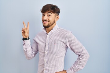 Wall Mural - Arab man with beard standing over blue background smiling looking to the camera showing fingers doing victory sign. number two.