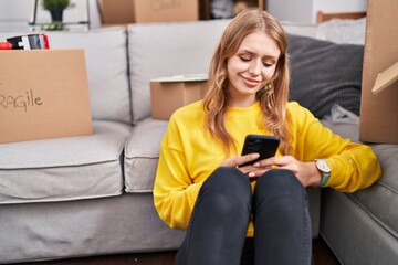 Wall Mural - Young blonde woman using smartphone sitting on floor at new home