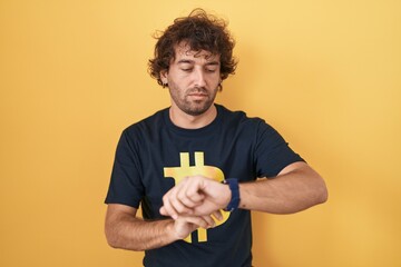 Poster - Hispanic young man wearing bitcoin t shirt checking the time on wrist watch, relaxed and confident