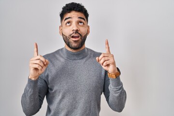 Wall Mural - Hispanic man with beard standing over white background amazed and surprised looking up and pointing with fingers and raised arms.