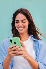 Vertical Portrait Of Smiling Happy Teenage Woman Using Her Smartphone And Chatting Online ,or Browsing Internet At Blue Teal Wall Background. Young Student Lady Watching Funny Videos With Mobile Phone
