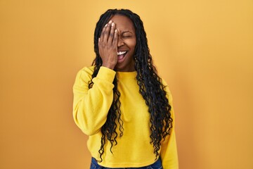 Poster - African woman standing over yellow background covering one eye with hand, confident smile on face and surprise emotion.