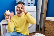 Hispanic Man Doing Laundry Holding Piggy Bank Smiling Happy Doing Ok Sign With Hand On Eye Looking Through Fingers