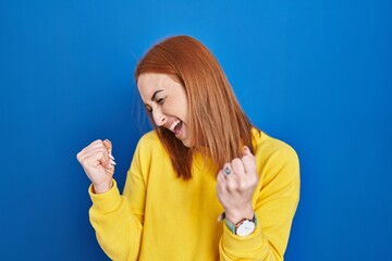 Wall Mural - Young woman standing over blue background very happy and excited doing winner gesture with arms raised, smiling and screaming for success. celebration concept.