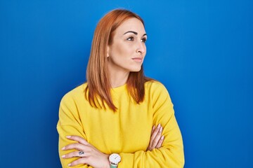 Sticker - Young woman standing over blue background looking to the side with arms crossed convinced and confident