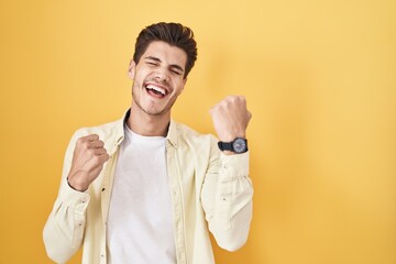 Wall Mural - Young hispanic man standing over yellow background very happy and excited doing winner gesture with arms raised, smiling and screaming for success. celebration concept.