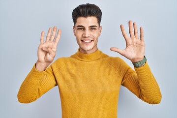 Wall Mural - Young hispanic man standing over blue background showing and pointing up with fingers number nine while smiling confident and happy.